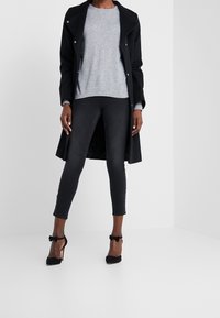 Un long manteau noir, un pull gris, des leggings noirs et des escarpins noirs avec un détail en nœud. Textures lisses, silhouette ajustée, et look superposé.