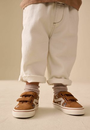 Braune Wildleder-Sneaker mit zwei Klettverschlüssen, weißen Akzenten und dekorativen Mustern. Kombiniert mit hochgekrempelten weißen Denim-Hosen und grauen Socken.