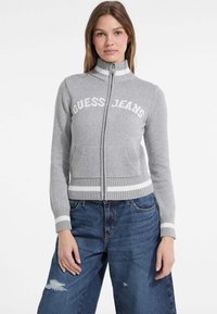 Šedý pletený svetrový zip s nápisom "GUESS JEANS", ribbovaným golierom a lemu, dvomi prednými vreckami a s bielymi pruhmi na manžetách a leme.