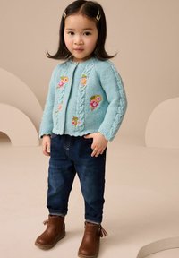 Cardigan lavorato a maglia di colore blu chiaro con ricami floreali, abbinato a jeans in denim scuro e stivaletti marroni alla caviglia. Il cardigan presenta dettagli a treccia.
