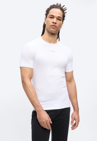 Άνδρας με dreadlocks που φοράει λευκό εφαρμοστό κοντομάνικο T-shirt και μαύρο παντελόνι, στέκεται μπροστά σε απλό ανοιχτό φόντο.