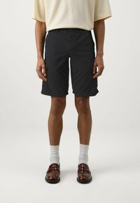 Pantaloni corti neri con una vestibilità rilassata, dotati di tasche laterali e chiusura con bottone. Indossati con calzini bianchi e mocassini marroni. Design semplice.