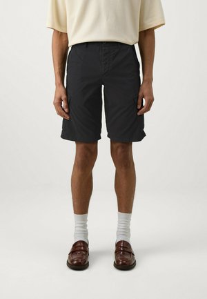 Pantaloni corti neri con una vestibilità rilassata, dotati di tasche laterali e chiusura con bottone. Indossati con calzini bianchi e mocassini marroni. Design semplice.