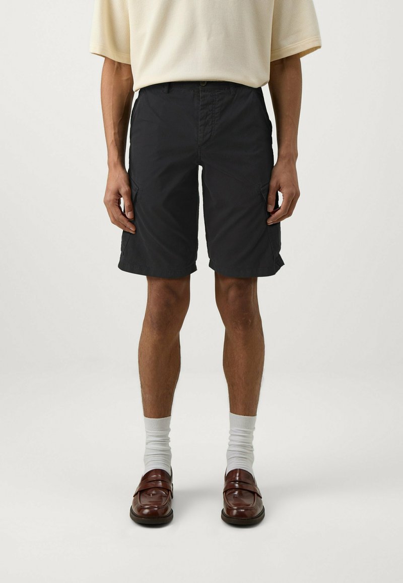 Pantaloni corti neri con una vestibilità rilassata, dotati di tasche laterali e chiusura con bottone. Indossati con calzini bianchi e mocassini marroni. Design semplice.