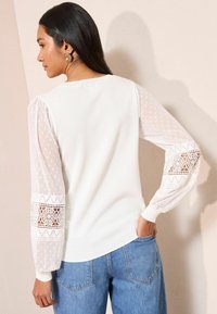 Fehér pulóver texturált csipkés lufi ujjakkal, pöttyös mintával és részletes csipke díszítéssel az ujjak végén. Kék, magasderekú farmerrel párosítva.