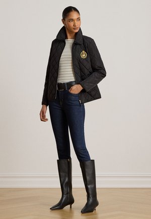 Kvinde iført en sort quiltet jakke med emblem, stribet skjorte, mørke jeans, sort bælte og sorte knælange støvler med hæl, stående indendørs.