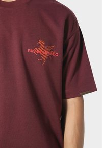 Musta t-paita, jossa on oranssiraidallinen hevosgrafiikka ja "PAS DE MONACO" -teksti rinnan kohdalla, lyhyet hihat näkyvät käsivarressa.