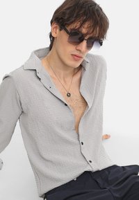 Camisa texturizada de color gris claro con diseño de botones, botones negros y cuello. Llevada con pantalones oscuros y gafas de sol, con un collar simple.