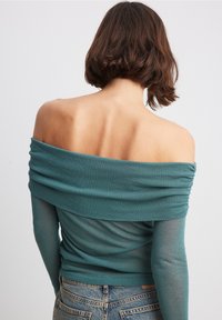 Tiefgrünes Carmen-Oberteil mit gerafftem Detail am Kragen, langen transparenten Ärmeln und gerafftem Körper, kombiniert mit Jeans.