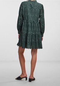 Dunkelgrünes Kleid mit langen Puffärmel, gestuften Saum und kunstvollen Spitzenverzierungen. Getragen mit schwarzen Absatzmules.