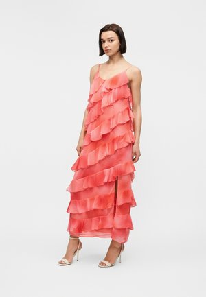 Woman wearing a sleeveless coral pink dress with layered ruffles and a front slit, paired with white high-heeled sandals.