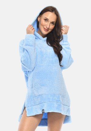 Frau trägt einen hellblauen, übergroßen, flauschigen Kapuzenpullover mit Kapuze, hält die Kapuze mit beiden Händen und lächelt sanft.