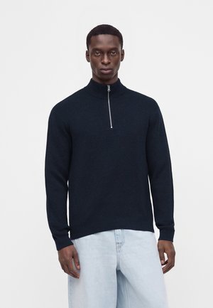 Homme portant un pull bleu marine foncé à demi-fermeture éclair et un jean bleu clair, debout devant un fond blanc uni.
