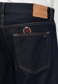 Mørke denimjeans har et lærmerke i livet og et brodert emblem på baklommen, som fremhever håndverk og detaljer.
