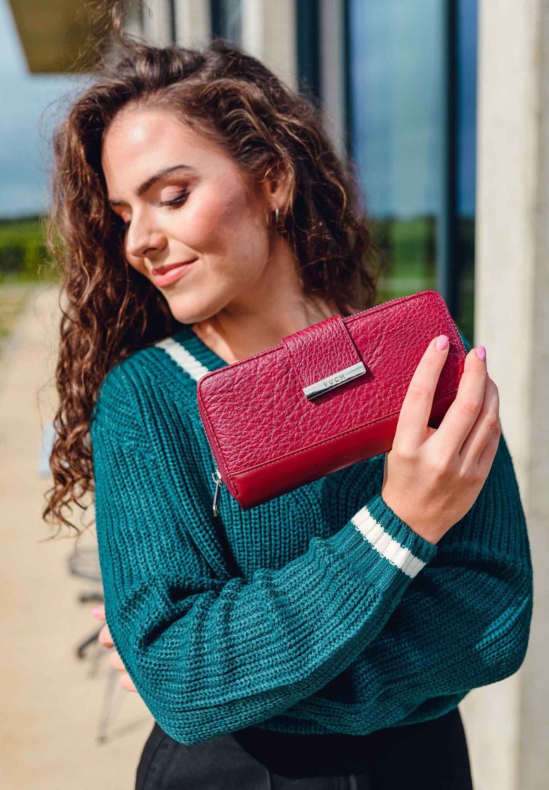 Red leather wallet with a textured surface, featuring a silver clasp and zipper. Handheld against a green sweater with white accents.