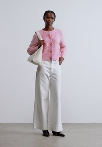 Rózsaszín, kötött kardigán arany gombokkal, fehér bő szárú nadrággal és fekete bokacsizmával párosítva. Egy fehér táskát tart a kezében. Egyszínű háttér.