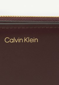 Portofel din piele burgundă închisă, cu fermoar auriu și logo-ul embosat "Calvin Klein" pe față, având o suprafață texturată.