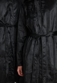Black satin long coats with textured surface, featuring belted waists and long sleeves, displayed side by side, partially obscured hands.
