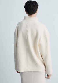 Crème wolblend oversized jas met hoge kraag en verlaagde schouders; zachte textuur en zichtbare naden op de rug.