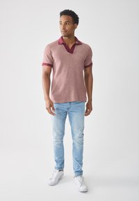 Hombre de pie con una polo de manga corta rosa, jeans azul claros y zapatillas blancas contra un fondo clarito, mirando ligeramente a la derecha.