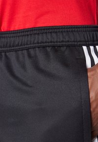 Schwarze Sportshorts mit elastischem Bund, ausgestattet mit einer Seitentasche und drei weißen Streifen entlang der linken Seite. Strukturierter Stoff.
