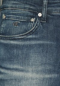 Primo piano di una tasca di jeans blu in denim con rivetti argentati e una piccola tasca cucita caratterizzata da ricamo del logo "cK".