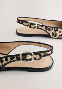 Schwarze flache Sandalen mit beigen und schwarzen, leopardenbedruckten geflochtenen Riemen und goldenen Schnallenverschlüssen vor hellem Hintergrund.