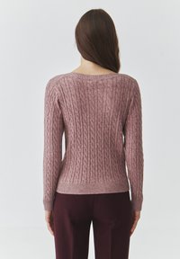 Cable-knit sweater in soft pink, featuring a wide neckline and ribbed hem. Paired with dark burgundy trousers, showing a fitted silhouette.