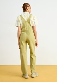 Salopette di cotone verde con vestibilità ampia, dotata di spalline regolabili, tasche posteriori e design a gamba dritta. Indossata sopra una maglietta a righe.