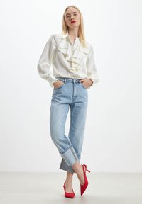 White ruffled blouse with long sleeves, light blue high-waisted jeans, and red pointed-toe heels, set against a plain background.