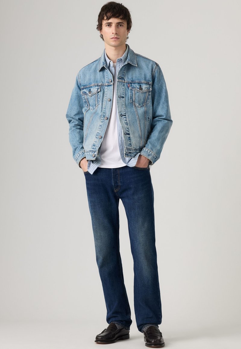 Young man standing with hands in pockets wearing a light blue denim jacket, white t-shirt, dark blue jeans, and black shoes.