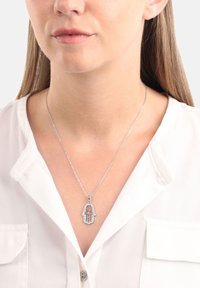 Silbernes Halsband mit einem Hamsa-Hand-Anhänger, der mit filigranen Details verziert ist. Über einem weißen Hemd getragen, ist die Kette zart und fein.