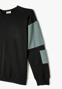 Svart sweatshirt med teal-färgade tygdetaljer på ärmarna. Har en ribbad halsringning och muddar; fickdetalj på vänster ärm. Mjuk textur.