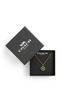 Pendentif circulaire vert avec contour doré, arborant un monogramme 'C'. Collier de chaîne à l'intérieur d'une boîte noire texturée avec un logo blanc.