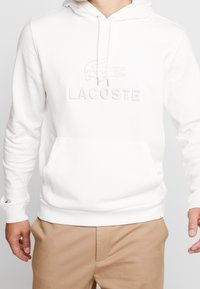 Vit hoodie med en framficka i kangurustil, med präglad "LACOSTE"-logotyp och krokodilsymbol. Tillverkad av mjukt, strukturerat tyg.