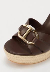 Brown leather wedge sandal with braided jute platform, featuring a gold buckle detail and soft straps. Smooth texture and neutral hue.