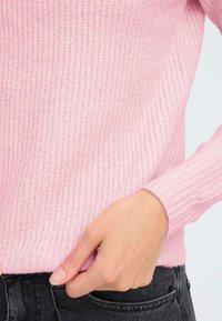 Pulover tricotat de culoare roz pal, cu o textură ribbed și manșete ajustate, purtat peste blugi închiși. Prim-plan al unei mâini care ajustează tivul.