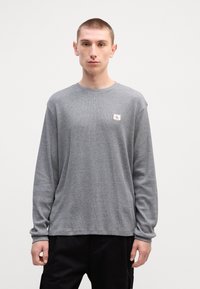 Γκρίζο μπλουζάκι μακρυμάνικο από υφασμάτινη ύφανση, με στρογγυλή λαιμόκοψη και μικρή ετικέτα λογότυπου στο αριστερό στήθος. Μαύρο παντελόνι.