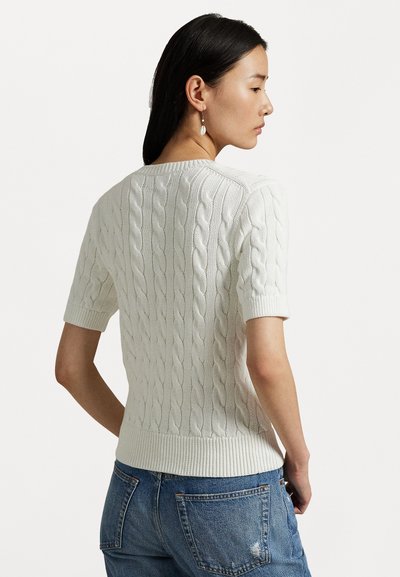 Pull à manches courtes en tricot torsadé crème avec des bords côtelés et un design texturé, associé à un jean en denim bleu. Vue de l'arrière.