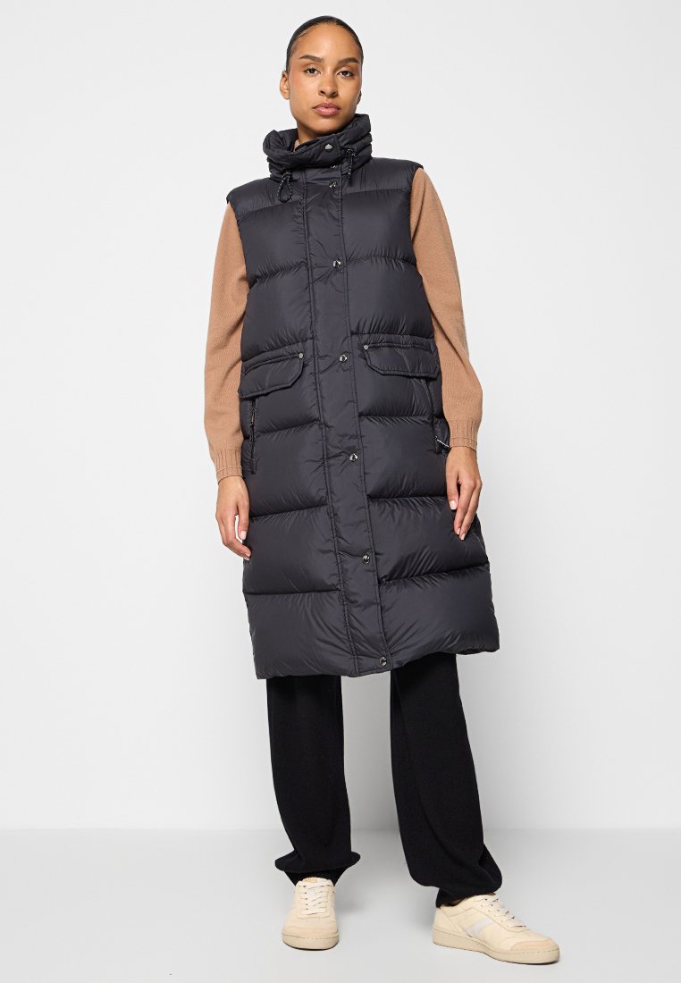 Langer schwarzer Pufferweste mit hohem Kragen, Druckknöpfen und zwei Fronttaschen, getragen über einem braunen Pullover und schwarzen Hosen. Hellfarbene Schuhe.