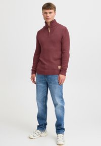 Burgunderfarbener Strickpullover mit hohem, geripptem Kragen und einem Holzreißverschluss. Kombiniert mit hellblauen Jeans und weißen Sneakers.
