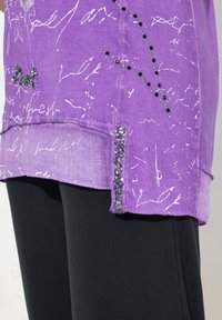 Paarse top met witte abstracte patronen; inclusief paillettenaccenten en een zoom met decoratieve details. Gecombineerd met zwarte broek.