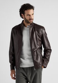 Brun läderjacka med ståkrage, zip-fickor och slät textur, samt en ljusgrå tröja och mörka byxor.