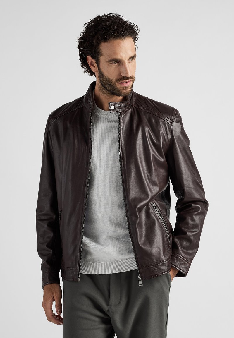 Brun läderjacka med ståkrage, zip-fickor och slät textur, samt en ljusgrå tröja och mörka byxor.