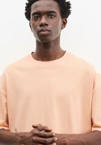 Lys ferskenfarvet oversized t-shirt med korte ærmer, rund halsudskæring, minimal syning detaljer og glat bomuldsstruktur. Modellens hænder er foldet.