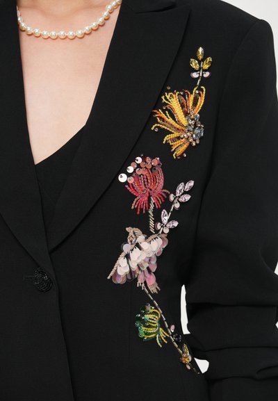 Fekete blézer élénk, színes virágos hímzéssel, texturált flitterekkel és díszítésekkel a bal oldalon.