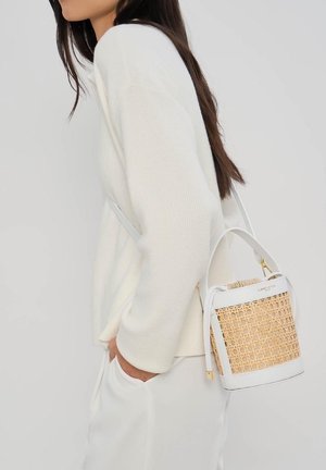 Femme en pull en maille blanc et pantalon portant un sac seau tissé blanc et naturel sur l'épaule.