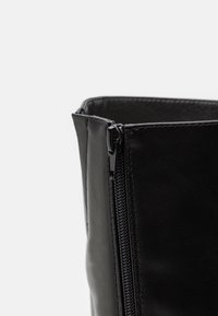 Bottine en cuir noir avec une fermeture éclair latérale, une texture lisse et des coutures renforcées. L'ouverture présente un bord légèrement incliné.