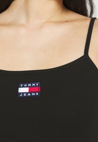 Czarny, prążkowany top na ramiączkach z prostokątną haftowaną naszywką w kolorze granatowym, czerwonym i białym z napisem "TOMMY JEANS". Cienkie ramiączka.
