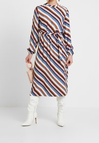 Gestreiftes Kleid mit langen Ärmeln, geraffter Taille und Bindegürtel. Enthält diagonale Streifen in Blau, Pink, Orange und Braun. Gestylt mit weißen, knielangen Stiefeln.